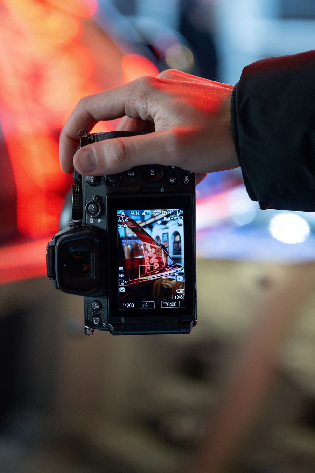 Appareil photo Nikon Z5II tenu à la main, affichant un bus rouge sur son écran, avec des lumières rouges floues en arrière-plan