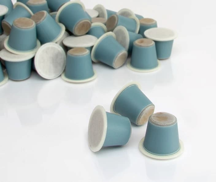 Capsules de café compostables gebana de couleur bleu-gris avec rebord blanc, dispersées sur fond blanc