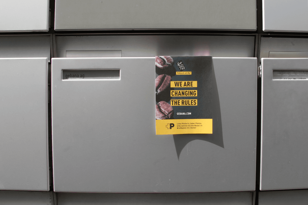 Affiche gebana collée sur une boîte aux lettres grise, avec texte jaune « We are changing the rules » et images de cerises de café