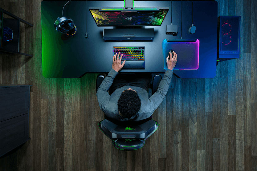 Dessus d'un gamer assis à son bureau gaming Razer avec écran, clavier RGB et accessoires illuminés en vert et bleu