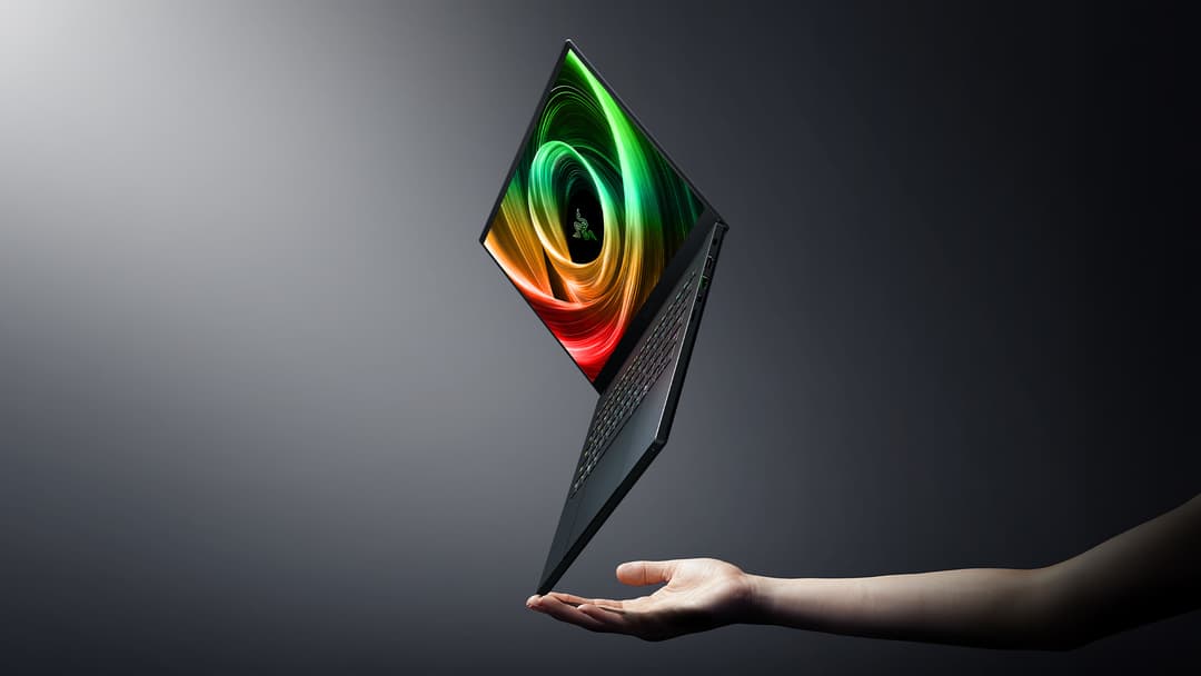 Razer Blade 14 ouvert en angle, tenu à la main, écran affichant un dégradé vert, orange et rouge, finition noire mate