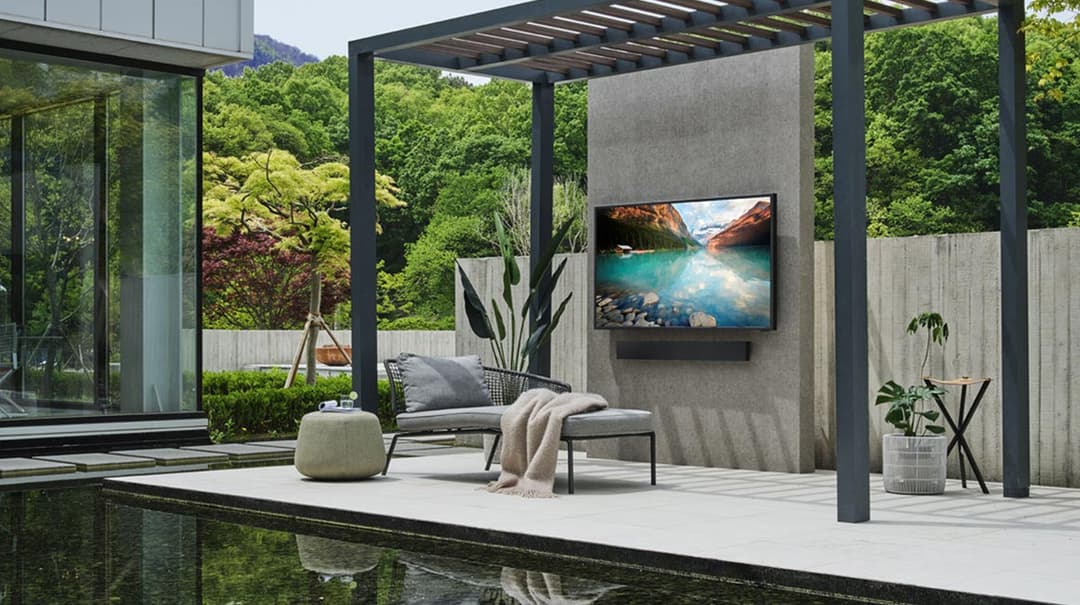 Téléviseur Samsung The Terrace intégré dans un mur gris d'une terrasse extérieure moderne avec pergola, mobilier de détente et piscine