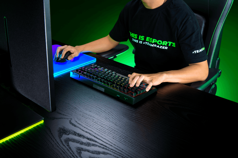 Joueur utilisant un clavier Razer BlackWidow V4 Tenkeyless avec souris bleue illuminée sur un bureau gaming vert et noir