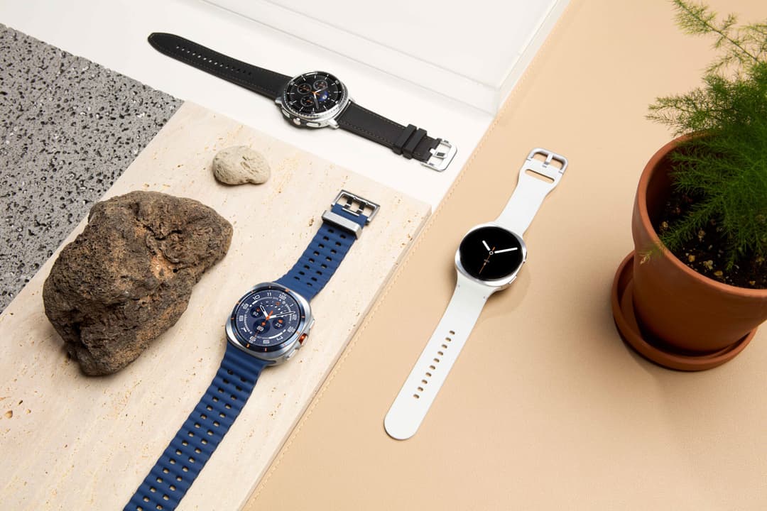 Trois montres Samsung Galaxy Watch Ultra et Watch8 de couleurs différentes, avec bracelets bleu, noir et blanc, disposées sur des surfaces texturées avec rochers et plante