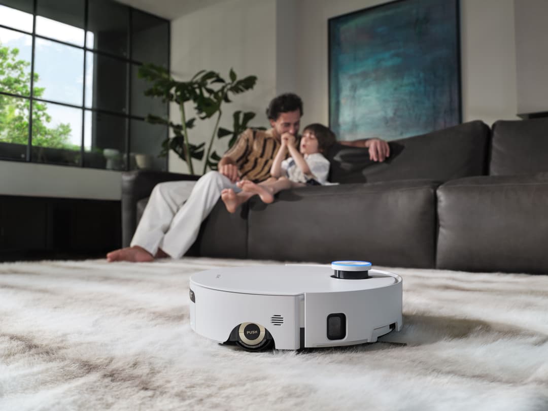 Robot aspirateur blanc Aqua10 Ultra Roller Complete en action sur un tapis, avec une famille assise sur un canapé gris en arrière-plan dans un salon moderne