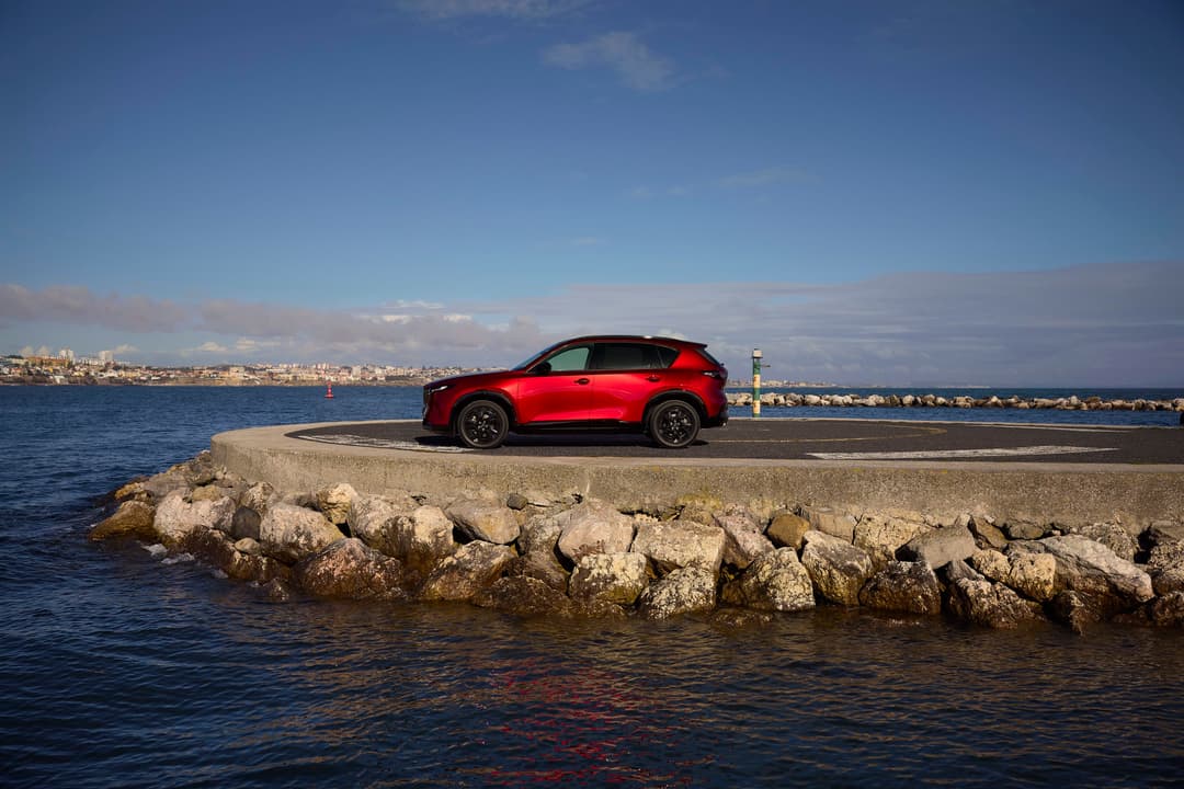 Nouveau Mazda CX-5 rouge garé sur une jetée en bord de mer, avec ville côtière en arrière-plan