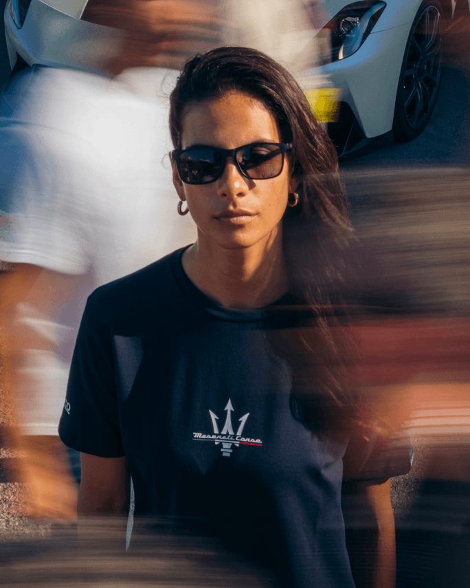 Femme portant un t-shirt Maserati Corse noir et des lunettes de soleil, devant une Maserati blanche en mouvement