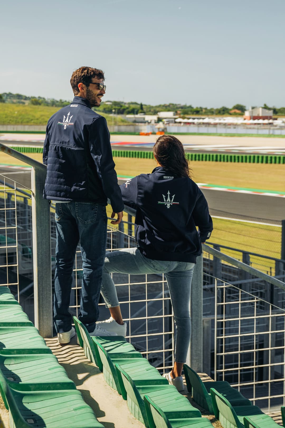 Deux personnes portant des vestes noires Maserati Sparco face à un circuit automobile vert et bleu