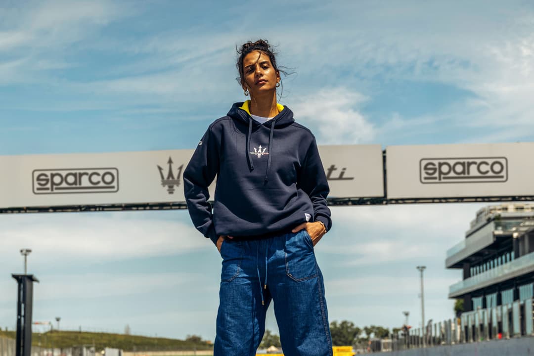 Femme portant un sweatshirt bleu marine Maserati Corse avec logo Sparco, sur un circuit automobile par beau temps