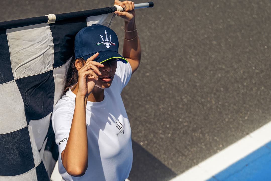 Personne tenant un drapeau Maserati Corse tout en portant une casquette et un t-shirt blanc de la collection Sparco X Maserati