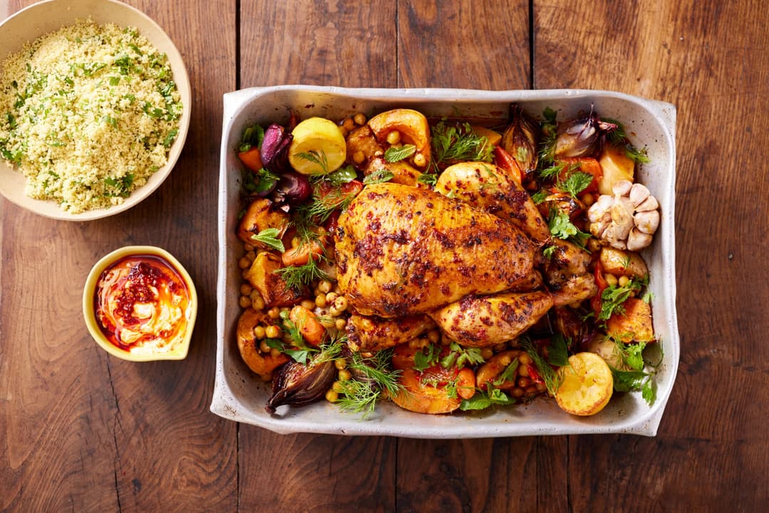 Poulet rôti garni de légumes colorés et d'herbes fraîches dans un plat blanc, accompagné d'une coupelle de sauce et d'un bol de couscous aux herbes