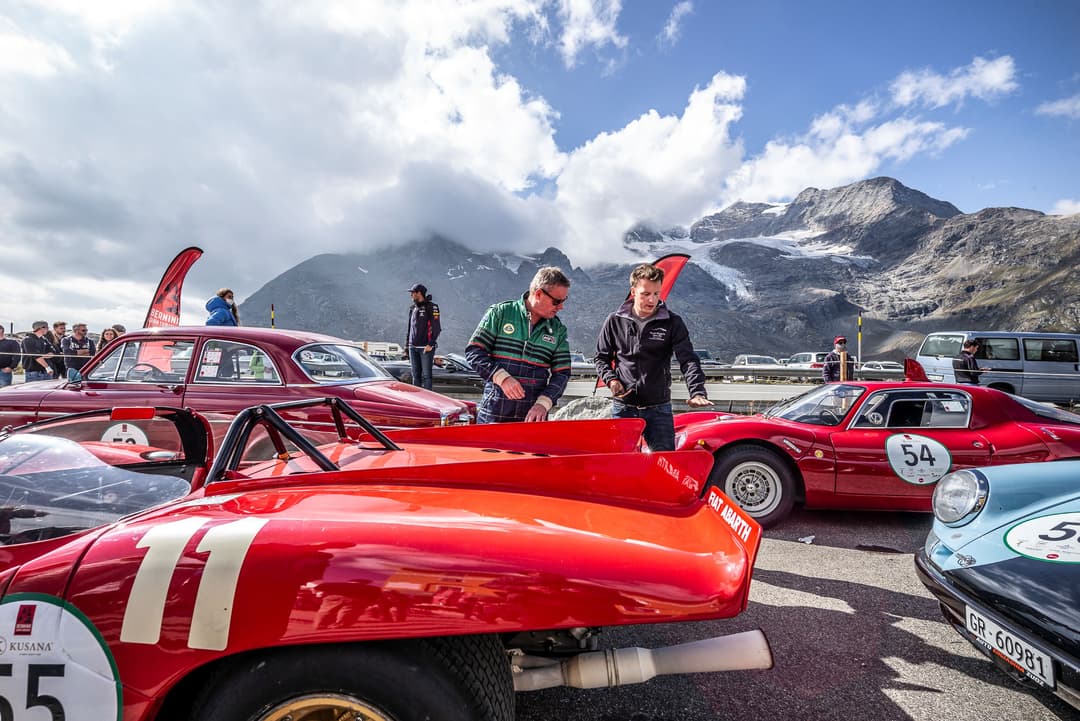 Voitures de course vintage rouges exposées en haute altitude, avec montagnes enneigées en arrière-plan et visiteurs admirant les véhicules