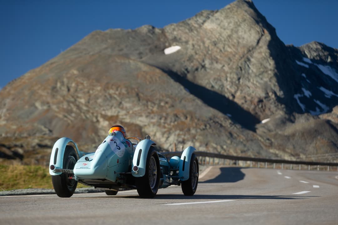 Voiture de course vintage blanche et bleue montant une route alpine vers des sommets rocheux sous un ciel dégagé