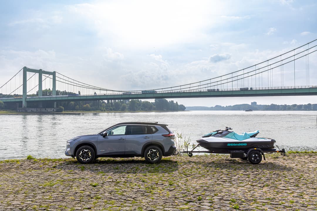 Nissan X-Trail gris garé au bord d'un fleuve avec un jet-ski tracté, grand pont suspendu en arrière-plan