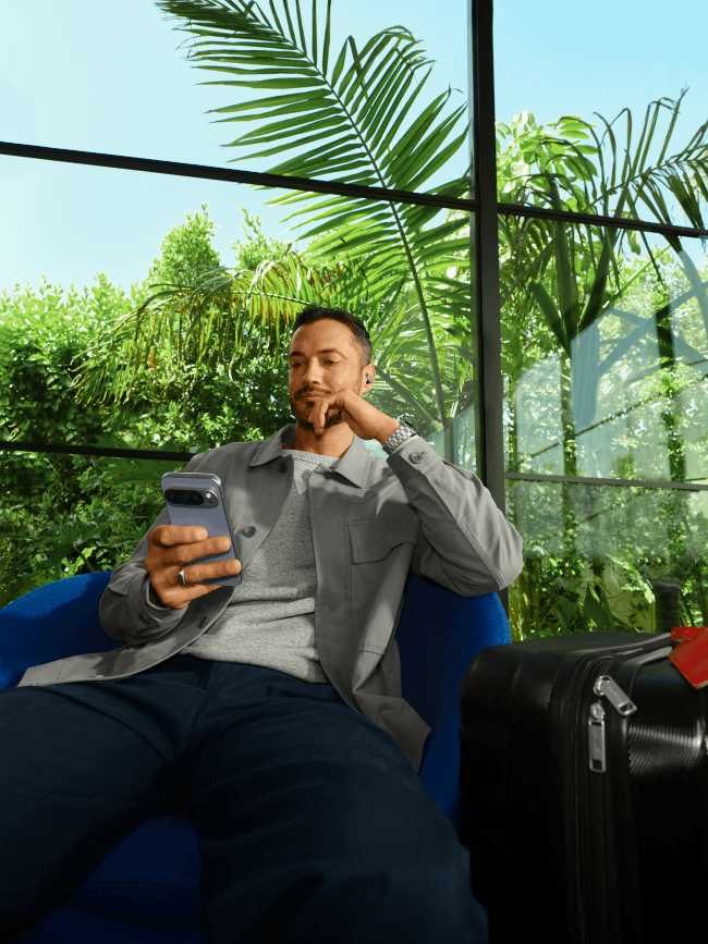 Homme tenant un smartphone Pixel 10 sous une véranda avec plantes vertes en arrière-plan, un appareil photo numérique à proximité