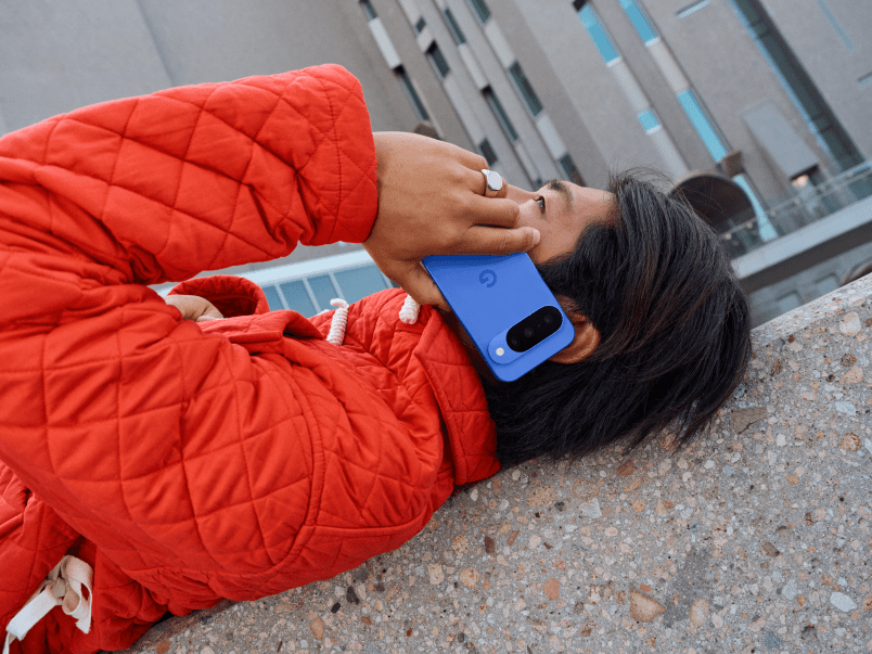 Personne en veste rouge tenant un Google Pixel 10 bleu avec sa caméra visible, photographiée de haut en milieu urbain