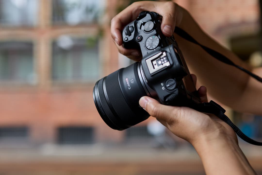 Mains tenant un appareil photo reflex Canon EOS R6 Mark III avec objectif RF 45mm, en extérieur devant un bâtiment en brique