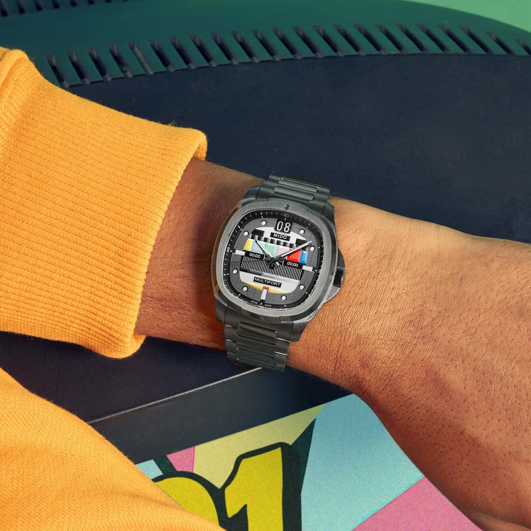 Montre Mido Multifort TV Big Date au boîtier gris, cadran noir avec éléments colorés, bracelet métallique acier, portée au poignet avec pull jaune