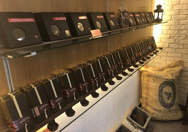 Intérieur de chocolaterie avec étagères en bois présentant des tablettes et des emballages de chocolat noir, sacs de jute de cacao visible à droite