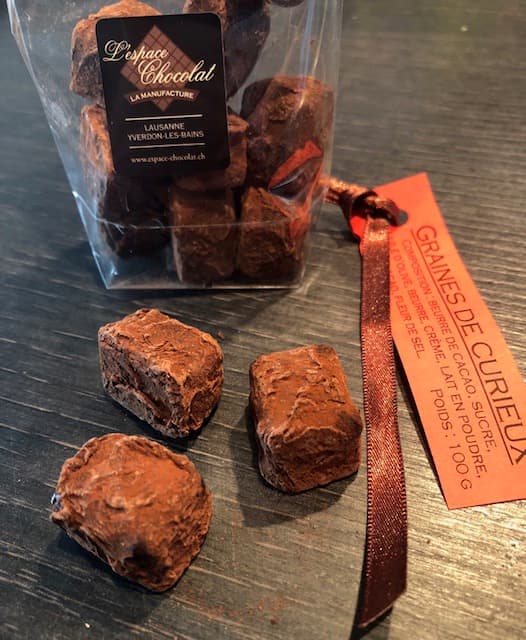 Graines de Curieux, bonbons chocolatés de l'Espace Chocolat, présentés à côté de leur emballage orange et d'un pot de chocolats