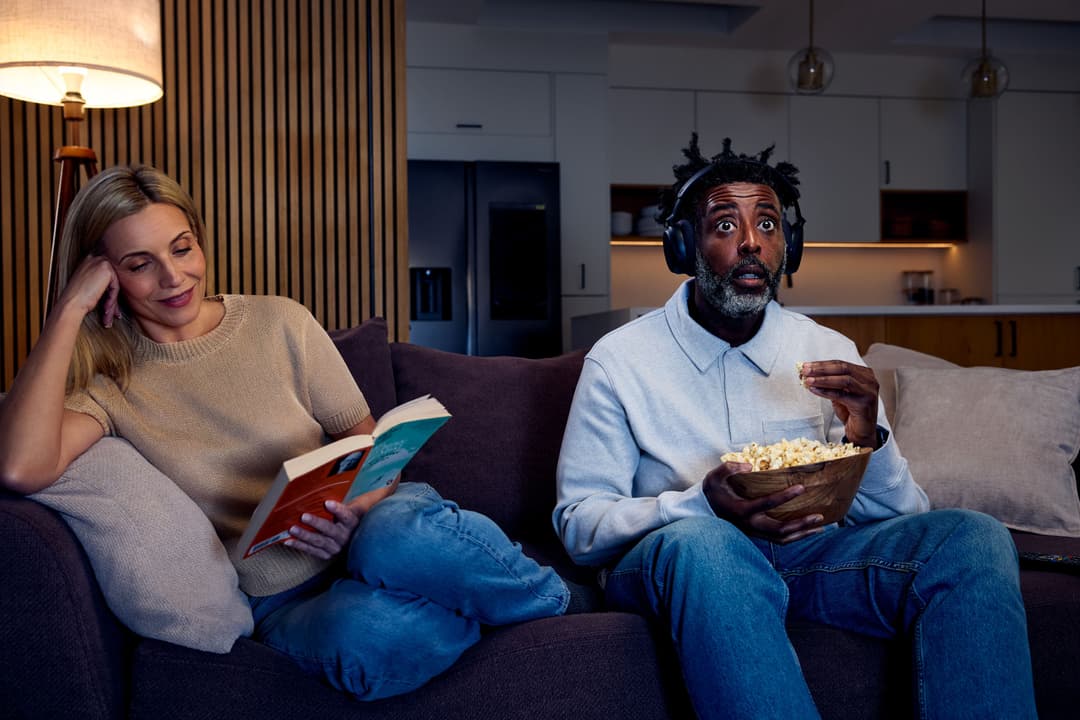 Deux personnes sur un canapé dans un salon moderne : l'une lit, l'autre regarde un écran avec des écouteurs et mange du pop-corn