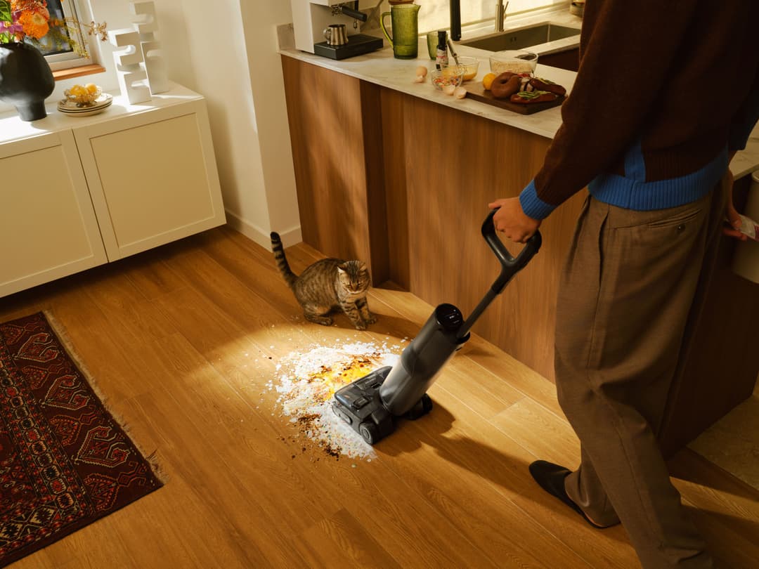 Roborock F25 Ace Pro aspirant des miettes sur un parquet en bois, manœuvré par une personne dans une cuisine moderne avec chat
