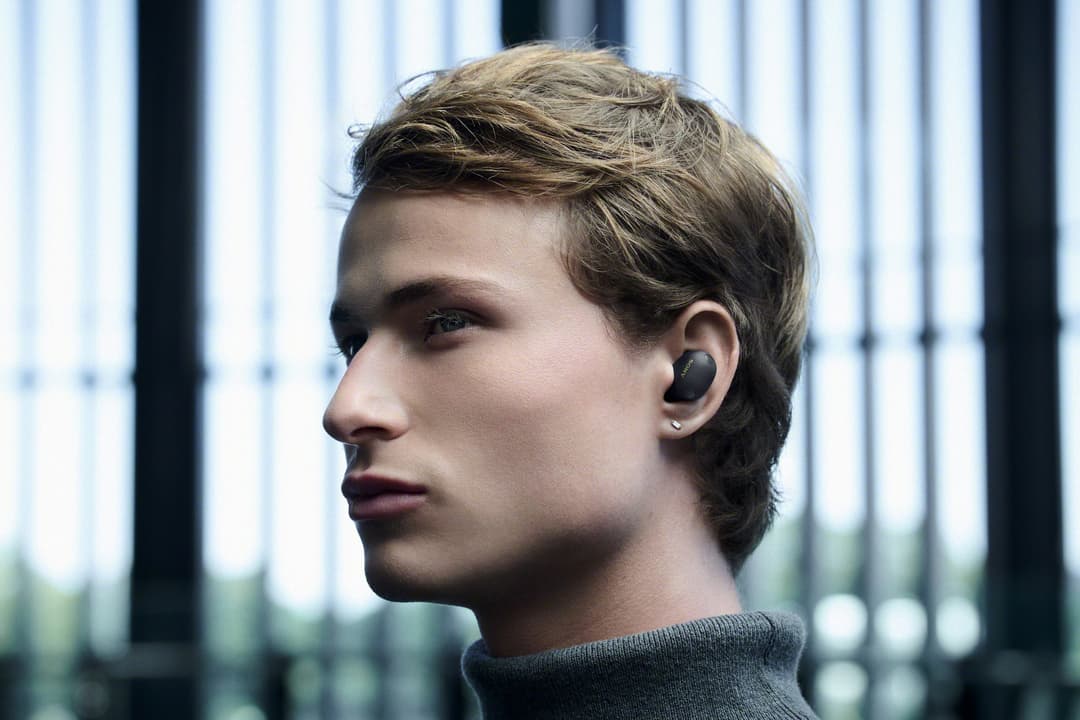 Homme de profil portant un écouteur sans fil noir Sony WF-1000XM6 à l'oreille, fond urbain flou