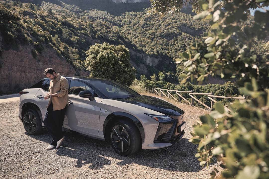 Lexus blanche avec un homme adossé à la portière, route sinueuse en montagne avec forêt dense et falaises rocheuses