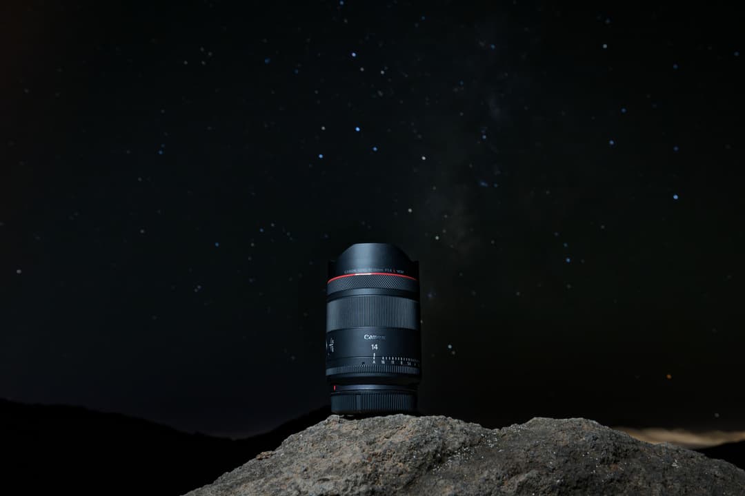 Objectif RF 14mm F1.4L Canon posé sur une pierre, ciel étoilé en arrière-plan