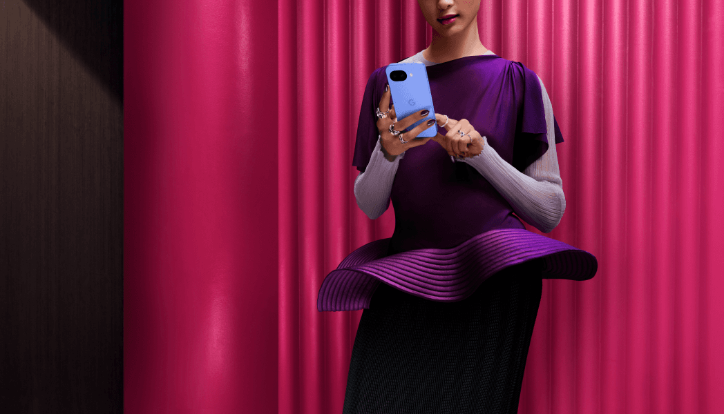 Google Pixel 10a bleu clair tenu à la main par une femme portant des vêtements noirs et violets, fond rose vif