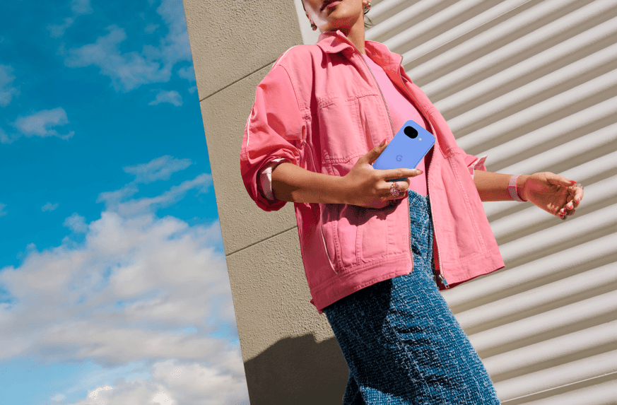 Personne tenant un Google Pixel 10a bleu clair dans sa main, vêtue d'une veste rose et d'un jean, devant un mur blanc avec ciel bleu en arrière-plan