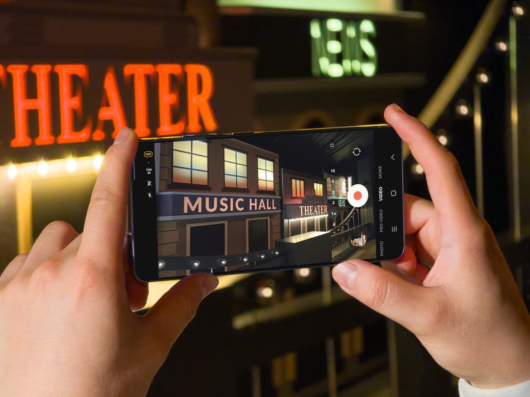 Mains tenant un Samsung Galaxy S26 filmant une façade de théâtre illuminée la nuit