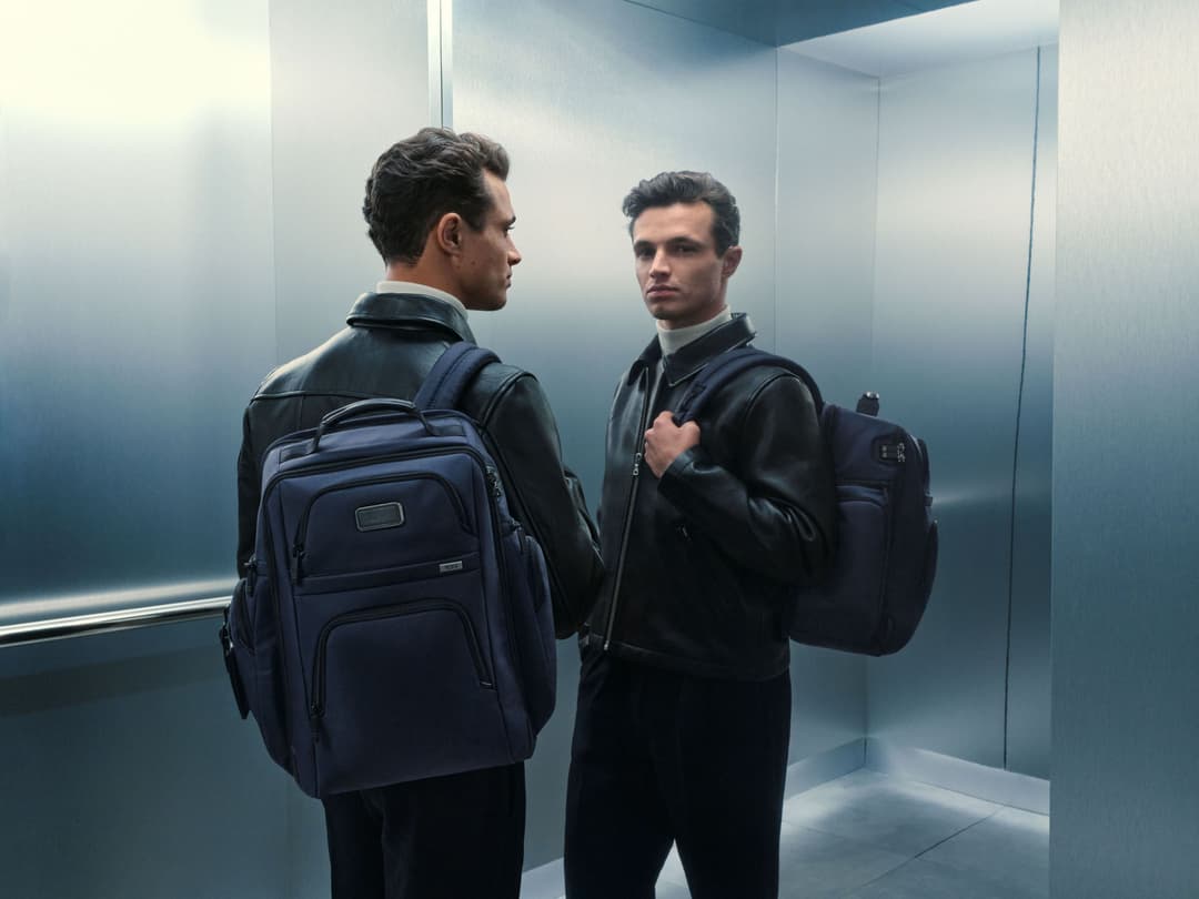 Lando Norris porte les bagages TUMI Alpha dans un décor minimaliste, vêtu d'une veste noire et de sacs de voyage bleu marine