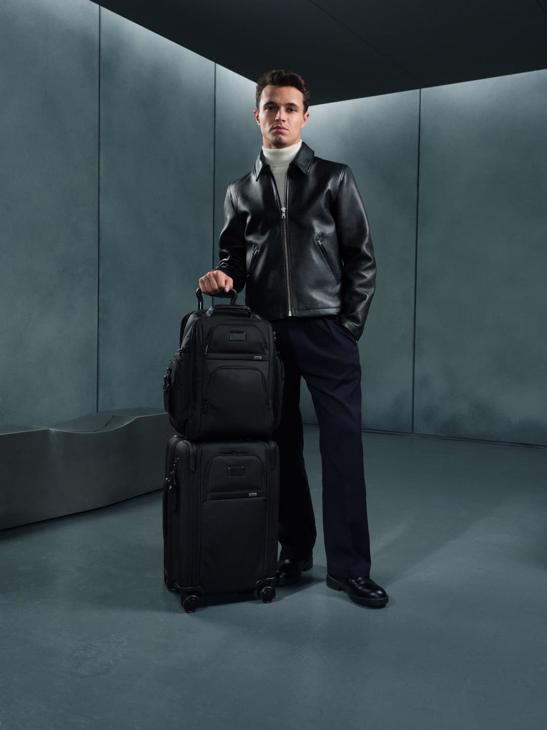 Lando Norris pose avec les bagages TUMI Alpha, veste de cuir noir sur fond architecturé gris bleuté