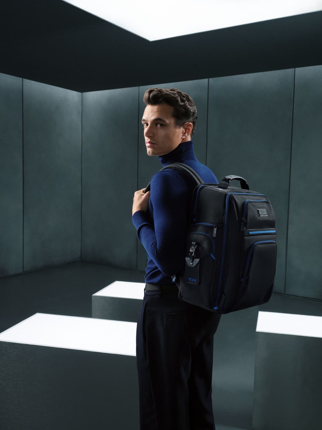 Homme portant un sac à dos TUMI noir avec accents bleus dans un intérieur épuré aux murs gris et ciel noir