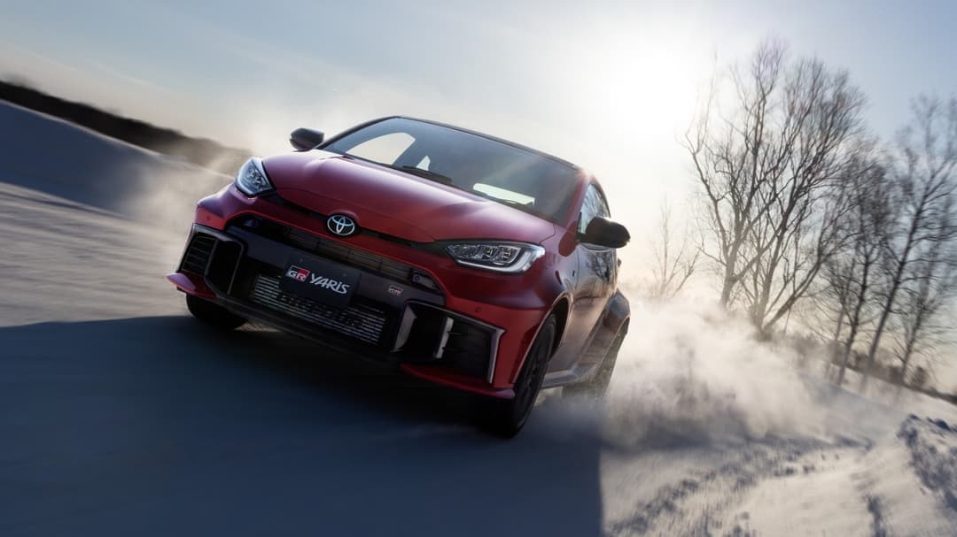 Toyota GR Yaris rouge en accélération sur piste enneigée, soulevant la poussière, avec arbres nus en arrière-plan