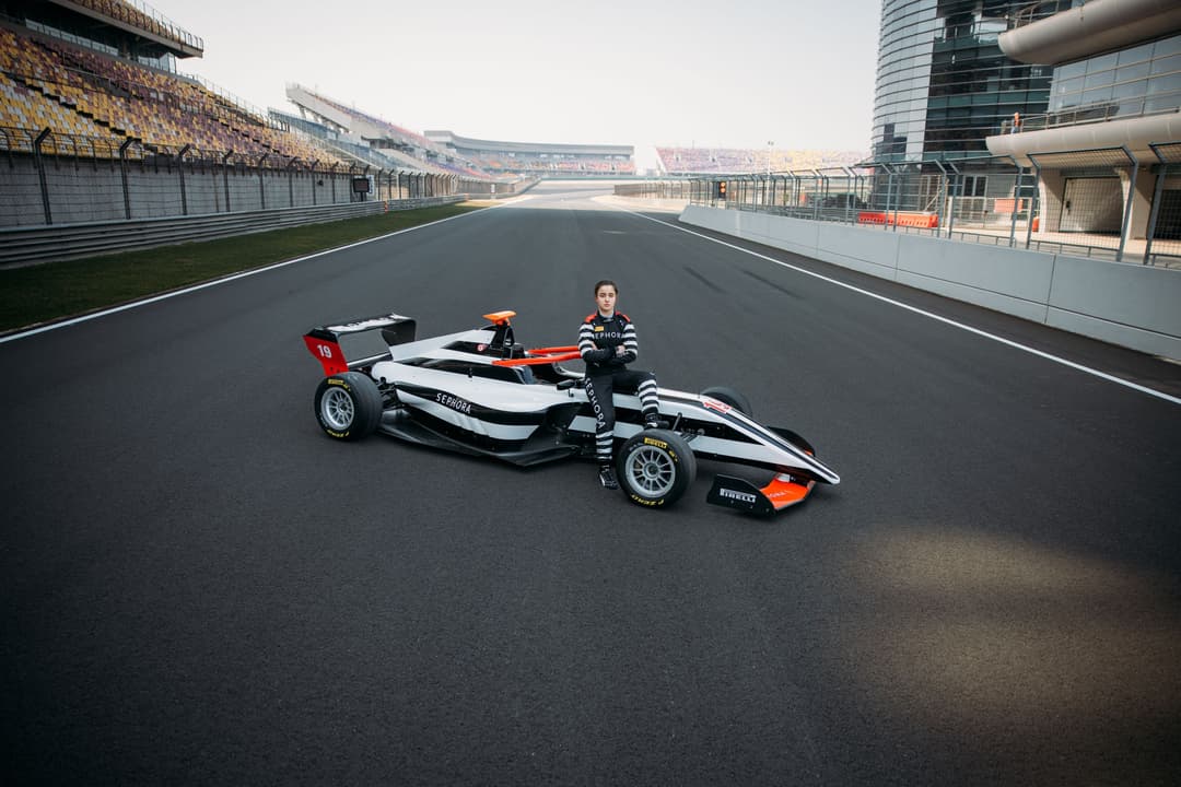 Pilote féminine posant à côté d'une monoplace F1 Academy rouge et blanche sur la piste d'un circuit de course
