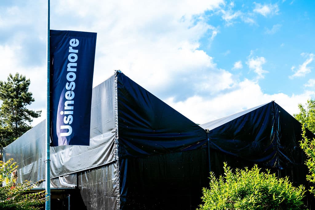 Bannière bleue et blanche du festival Usinesonore sur un mât, devant une structure couverte de bâches noires en plein air