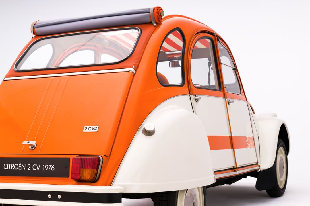 Citroën 2CV 1976 orange et blanche, profil trois-quarts arrière, avec galerie de toit et bandes latérales rouges