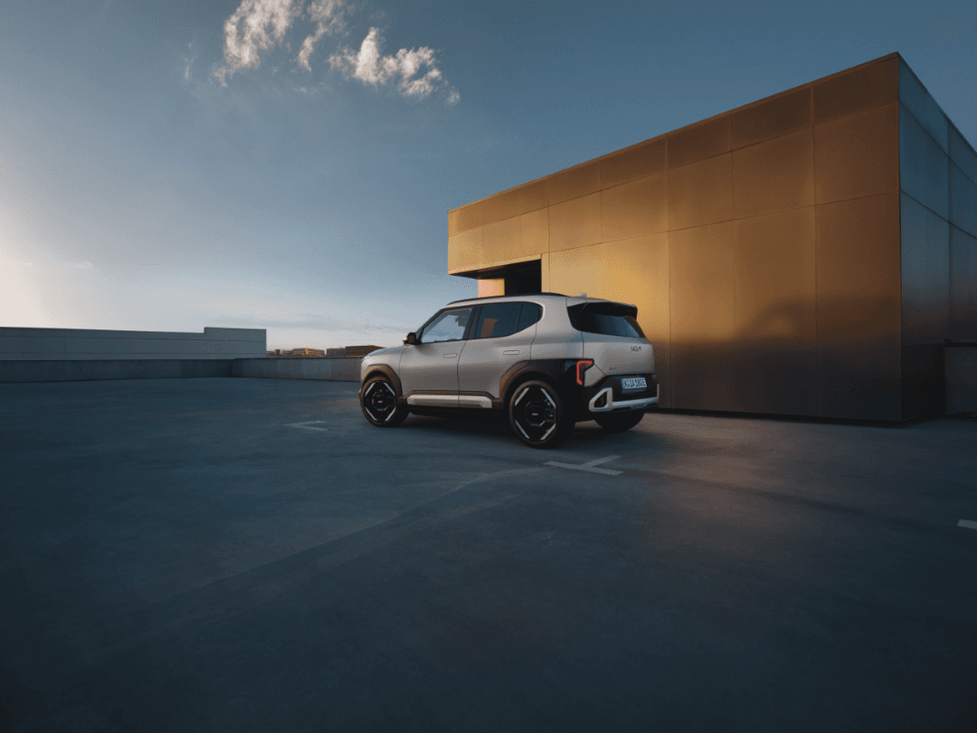 Kia EV2 blanc et noir, petit SUV électrique, garé sur une esplanade devant un bâtiment moderne aux façades dorées