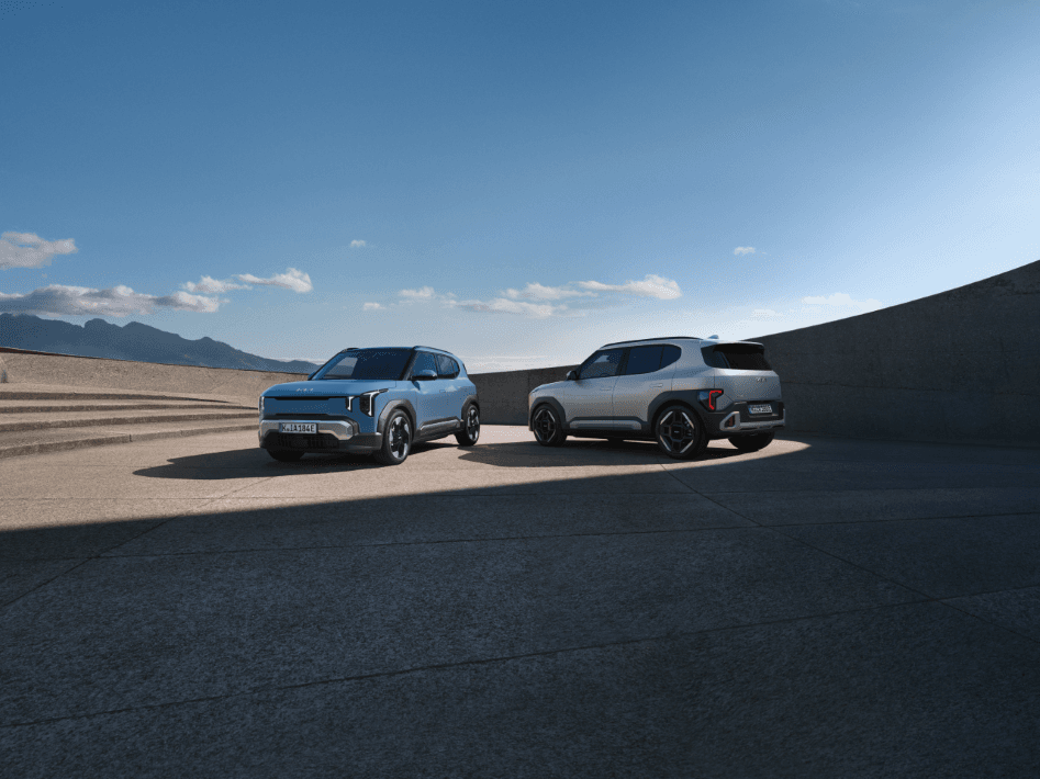 Deux Kia EV2 stationnés côte à côte sur un parking, l'un bleu ciel et l'autre gris argenté, avec montagne en arrière-plan