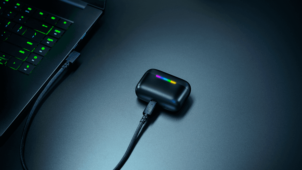 Boîtier de recharge noir des écouteurs Razer Hammerhead V3 HyperSpeed avec indicateurs LED colorés, connecté à un clavier gaming illuminé