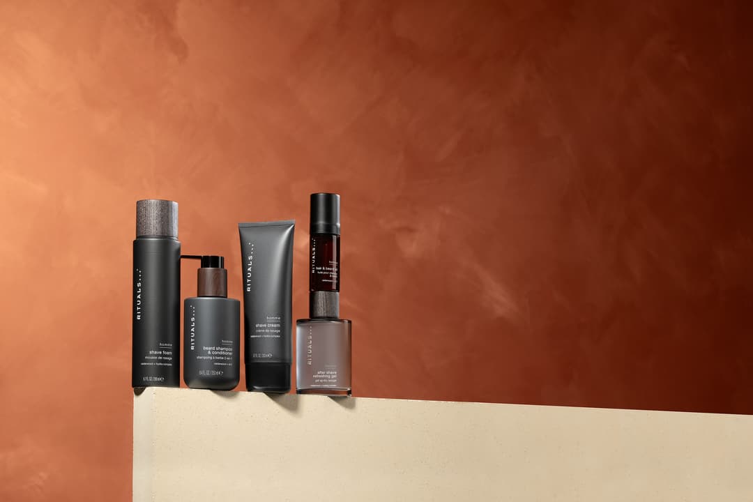 Cinq produits de soins Rituals Men Collection aux flacons noirs et teintés alignés sur fond beige et orange