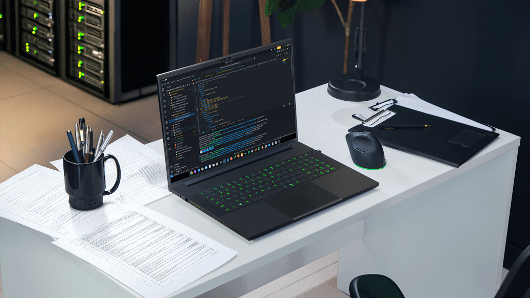 Laptop gamer Razer Blade 18 noir avec clavier rétroéclairé vert, écran affichant du code, posé sur un bureau blanc avec souris et accessoires