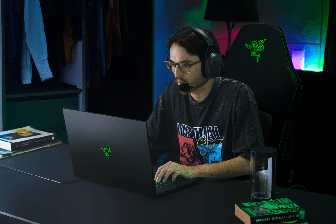 Homme portant des lunettes et un casque, assis à un bureau devant un laptop Razer Blade noir avec logo vert lumineux