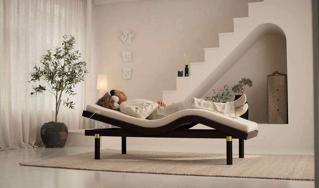Femme allongée sur un lit ergonomique blanc et noir dans un espace épuré beige avec plantes, escaliers et éclairage tamisé