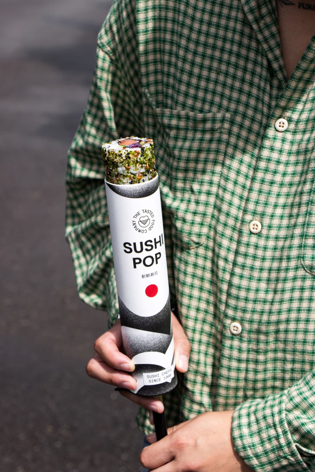 Sushi Pop en tube blanc et noir tenu à la main, sushi à la surface garnit d'œufs, personne portant chemise verte à carreaux