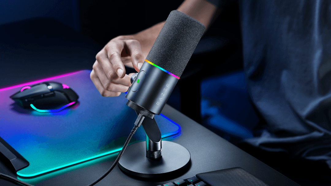 Microphone Seiren V3 Pro noir avec filtre anti-pop gris, tenu en main au-dessus de son support, sur un bureau gaming aux lumières bleues et roses