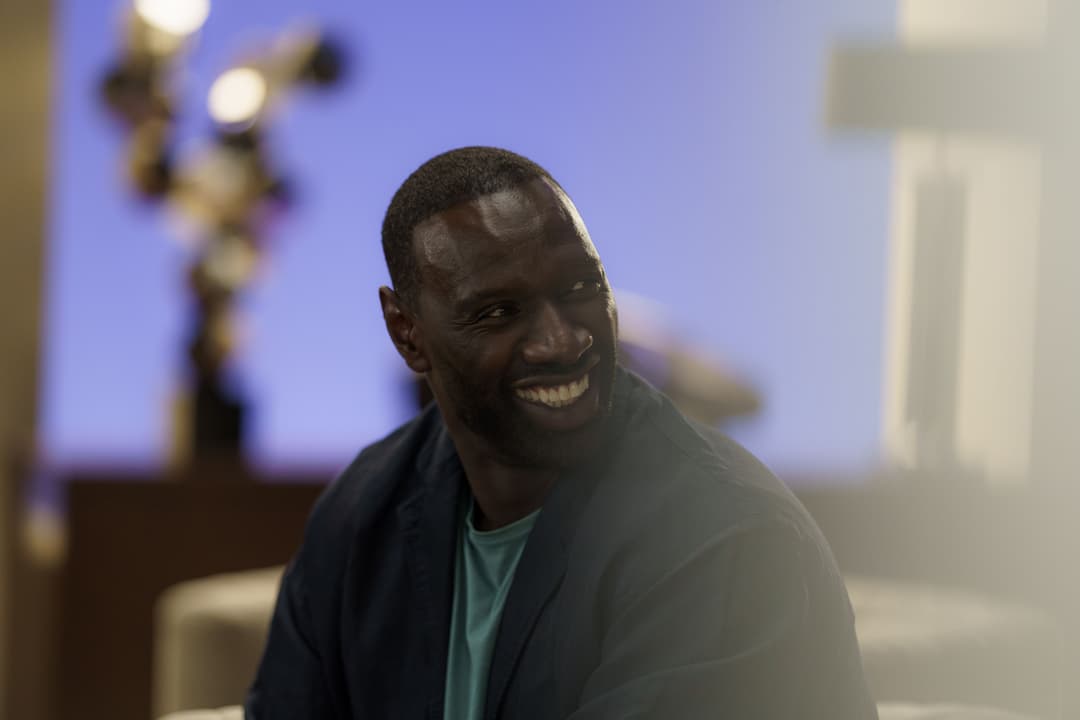 Homme souriant face à la caméra, vêtu d'une veste noire et d'un t-shirt vert, dans un intérieur moderne aux tons bleus