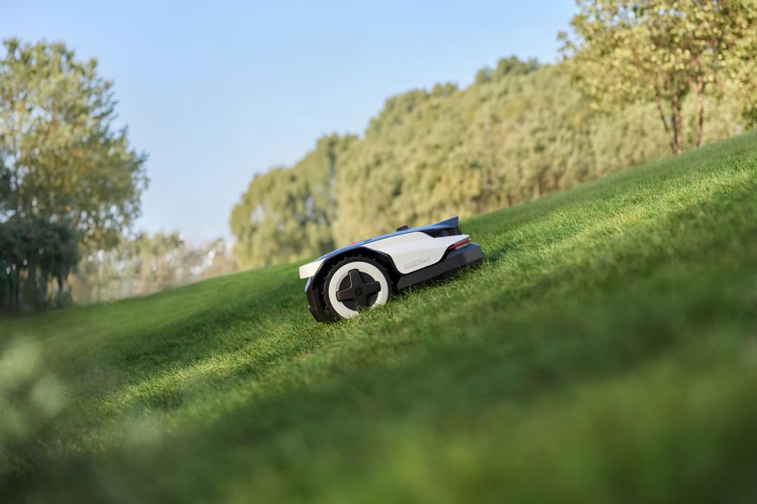 Robot tondeuse Roborock blanc et noir sur une pelouse en pente, avec haies et arbres en arrière-plan